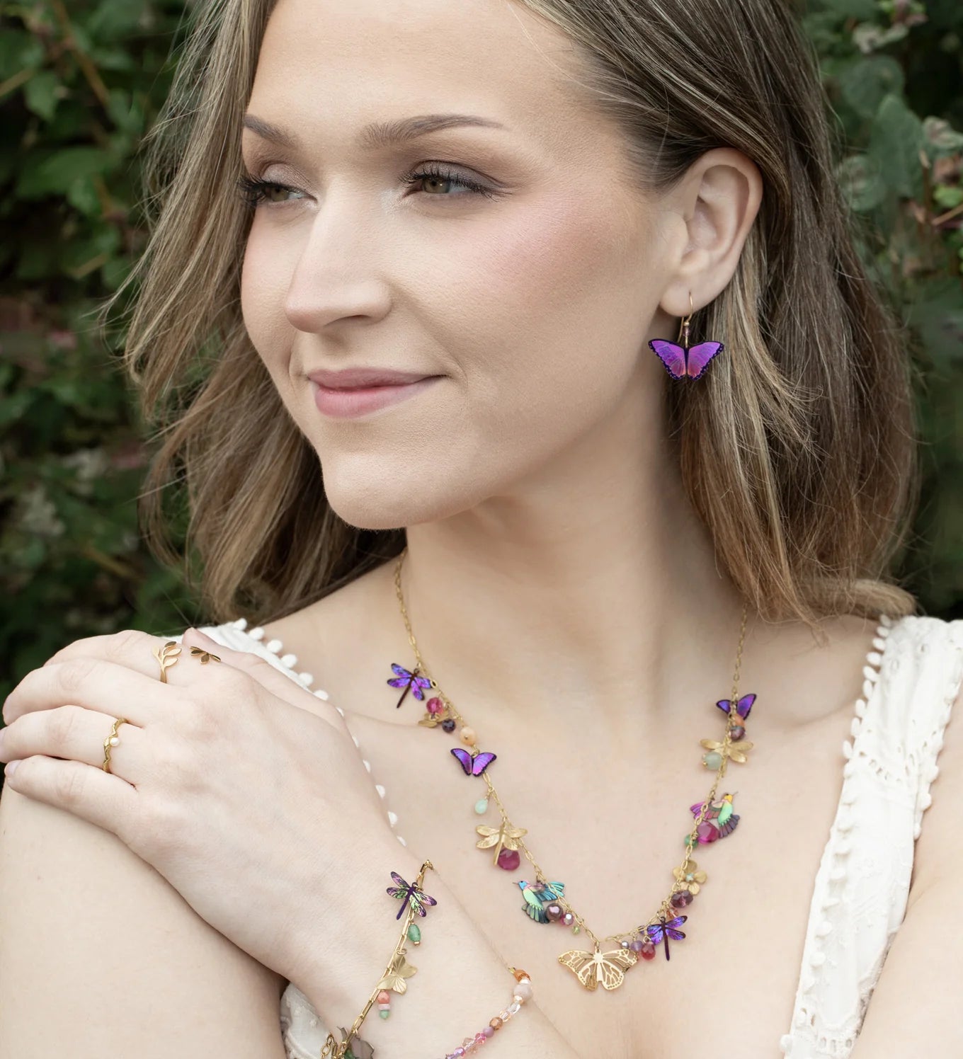 A woman with wavy blonde hair, smiling and looking to the side outdoors, wears a white top and colorful butterfly jewelry—including earrings, a necklace, and the Holly Yashi Garden Whimsy Gold Charms Bracelet adorned with gemstones.