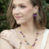 A woman with wavy blonde hair, smiling and looking to the side outdoors, wears a white top and colorful butterfly jewelry—including earrings, a necklace, and the Holly Yashi Garden Whimsy Gold Charms Bracelet adorned with gemstones.