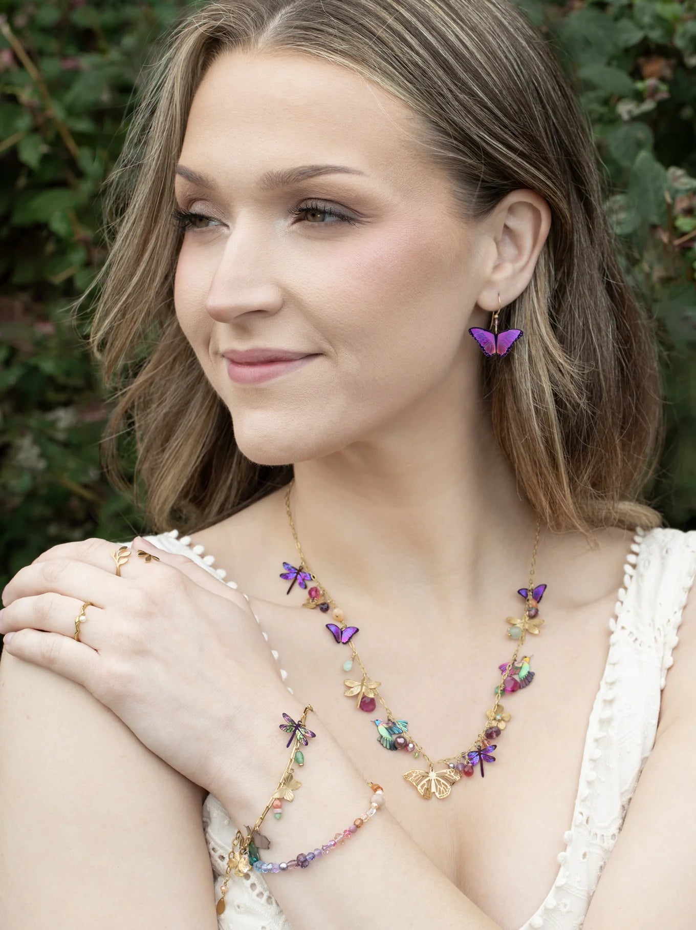 A woman with wavy blonde hair, smiling and looking to the side outdoors, wears a white top and colorful butterfly jewelry—including earrings, a necklace, and the Holly Yashi Garden Whimsy Gold Charms Bracelet adorned with gemstones.