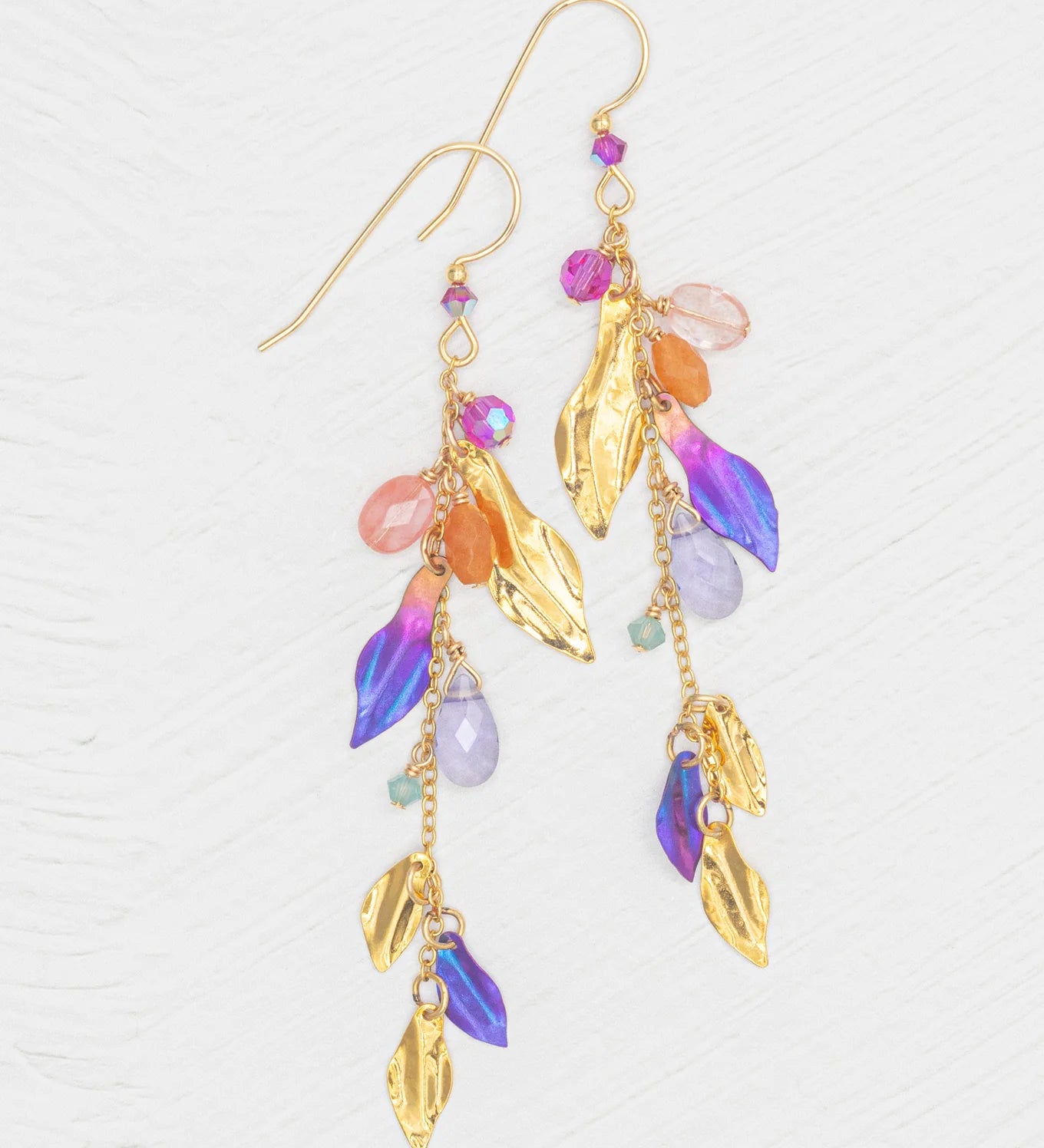 The Calliope Tropic Punch Earrings by Holly Yashi feature metallic gold leaves and vibrant purple, orange, pink, and turquoise beads. Hypoallergenic, they are elegantly displayed against a white textured background.
