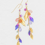 The Calliope Tropic Punch Earrings by Holly Yashi feature metallic gold leaves and vibrant purple, orange, pink, and turquoise beads. Hypoallergenic, they are elegantly displayed against a white textured background.