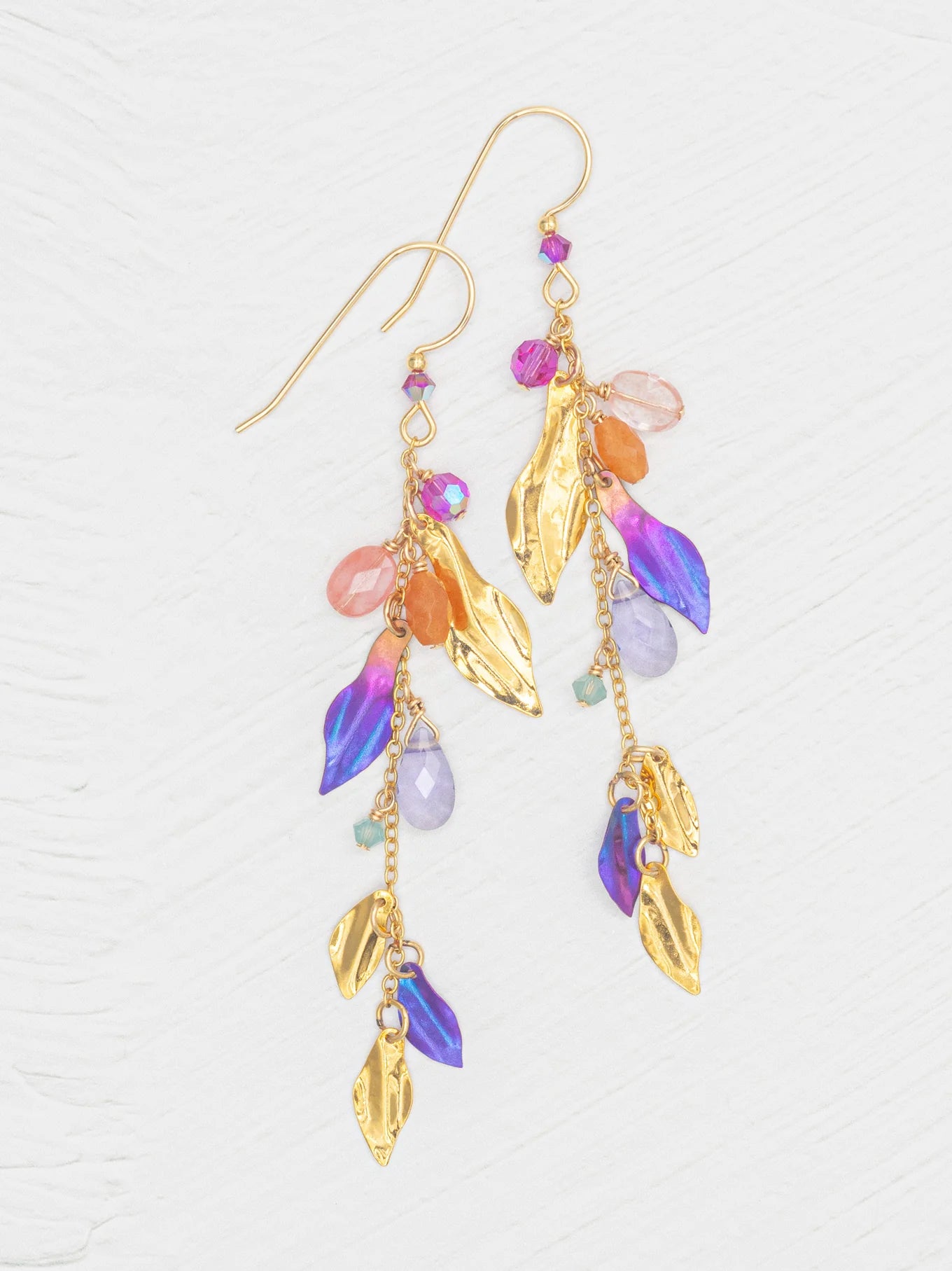 The Calliope Tropic Punch Earrings by Holly Yashi feature metallic gold leaves and vibrant purple, orange, pink, and turquoise beads. Hypoallergenic, they are elegantly displayed against a white textured background.