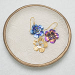 A textured leaf-patterned plate displays three Holly Yashi Blissful Bloom Gold & Silver Earrings, each with gold hooks, a central gemstone, and crafted hypo-allergenic for sensitive ears in blue, purple, and gold.