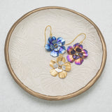 A textured leaf-patterned plate displays three Holly Yashi Blissful Bloom Gold & Silver Earrings, each with gold hooks, a central gemstone, and crafted hypo-allergenic for sensitive ears in blue, purple, and gold.
