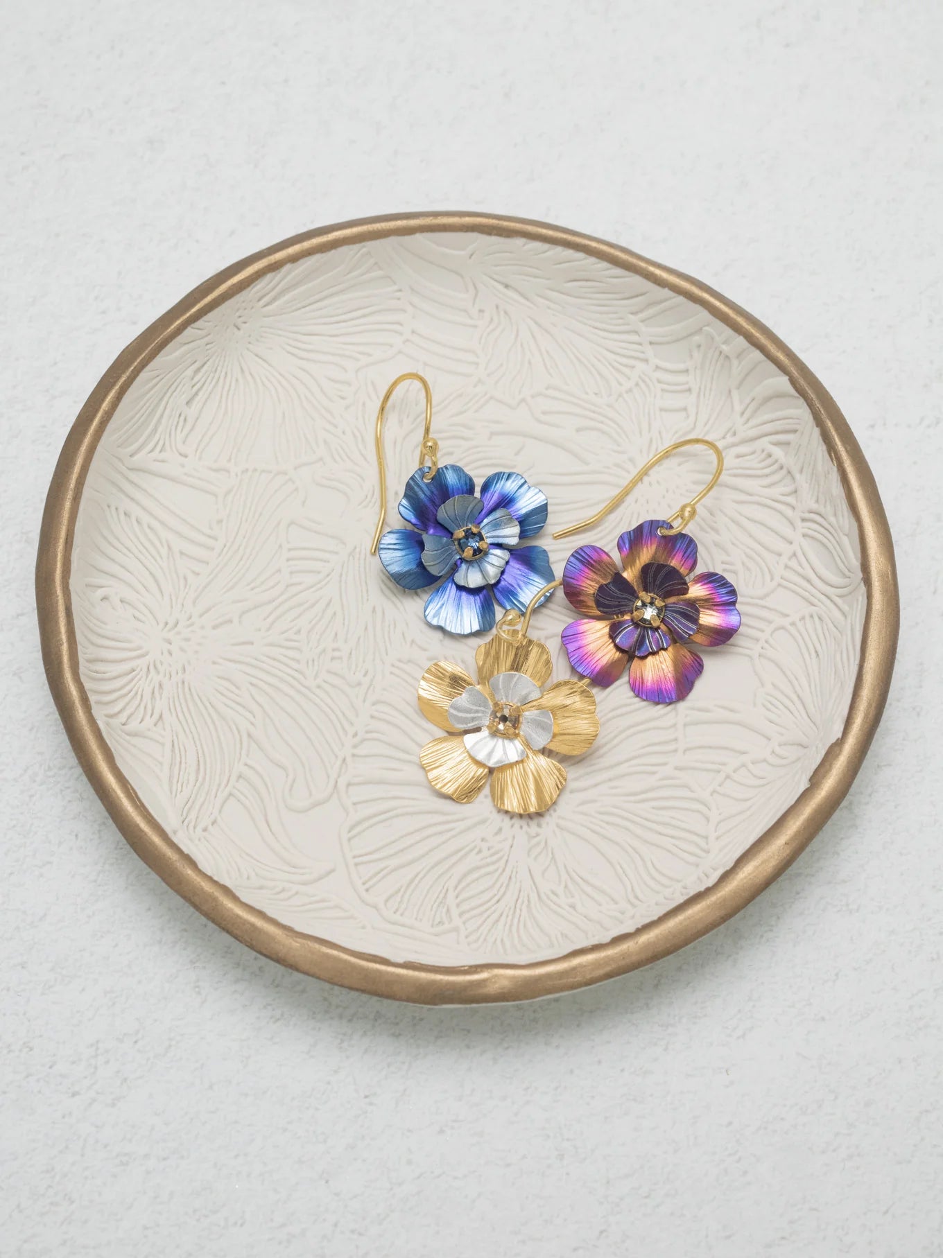 A textured leaf-patterned plate displays three Holly Yashi Blissful Bloom Gold & Silver Earrings, each with gold hooks, a central gemstone, and crafted hypo-allergenic for sensitive ears in blue, purple, and gold.
