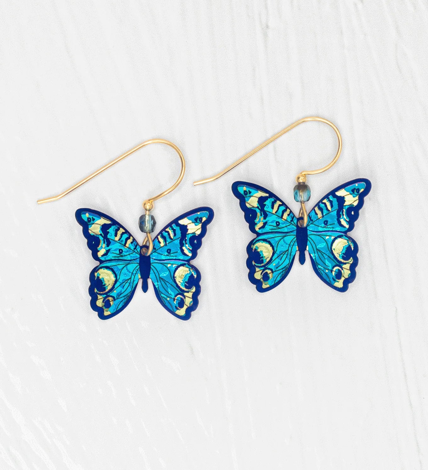The Holly Yashi ER: Mariposa Butterfly Blue Radiance earrings showcase blue butterfly charms with gold-filled accents and hooks, displayed on a white textured background. Hypoallergenic materials provide comfortable wear.