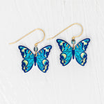 The Holly Yashi ER: Mariposa Butterfly Blue Radiance earrings showcase blue butterfly charms with gold-filled accents and hooks, displayed on a white textured background. Hypoallergenic materials provide comfortable wear.