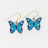 The Holly Yashi ER: Mariposa Butterfly Blue Radiance earrings showcase blue butterfly charms with gold-filled accents and hooks, displayed on a white textured background. Hypoallergenic materials provide comfortable wear.