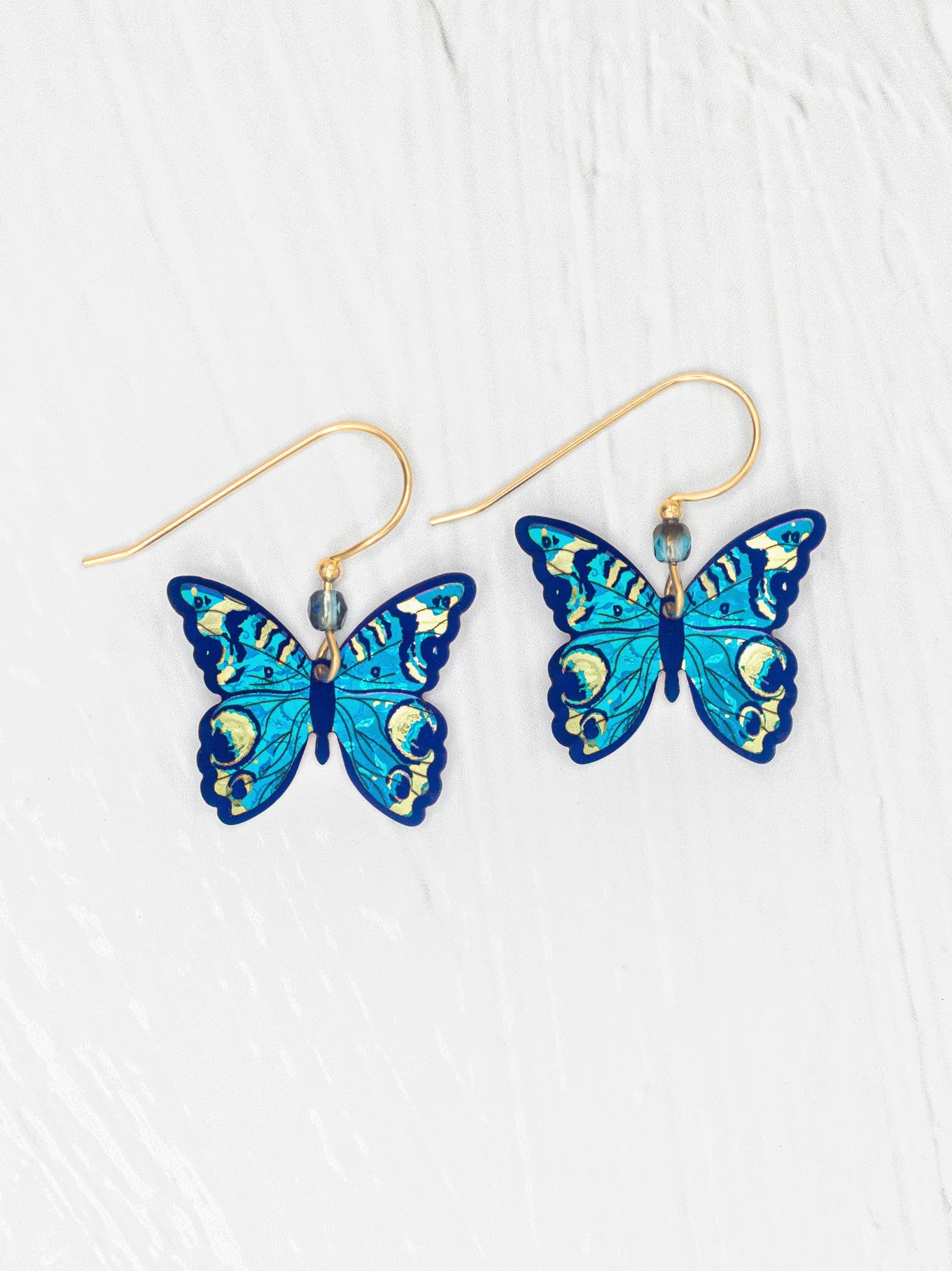 The Holly Yashi ER: Mariposa Butterfly Blue Radiance earrings showcase blue butterfly charms with gold-filled accents and hooks, displayed on a white textured background. Hypoallergenic materials provide comfortable wear.