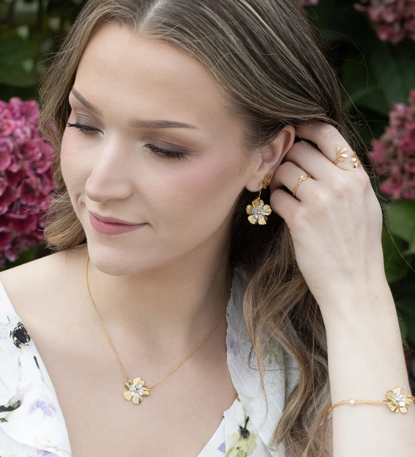 A woman with wavy brown hair wears a white floral dress and gold jewelry, including Holly Yashi’s Blissful Bloom Gold & Silver Earrings, while touching her hair and standing in front of pink hydrangea flowers.