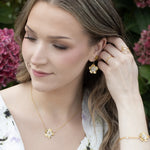 A woman with wavy brown hair wears a white floral dress and gold jewelry, including Holly Yashi’s Blissful Bloom Gold & Silver Earrings, while touching her hair and standing in front of pink hydrangea flowers.