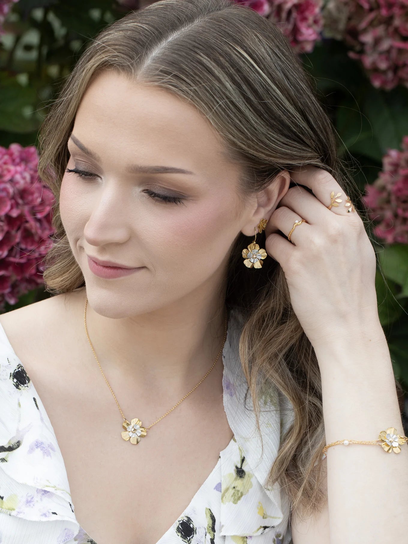 A woman with wavy brown hair wears a white floral dress and gold jewelry, including Holly Yashi’s Blissful Bloom Gold & Silver Earrings, while touching her hair and standing in front of pink hydrangea flowers.