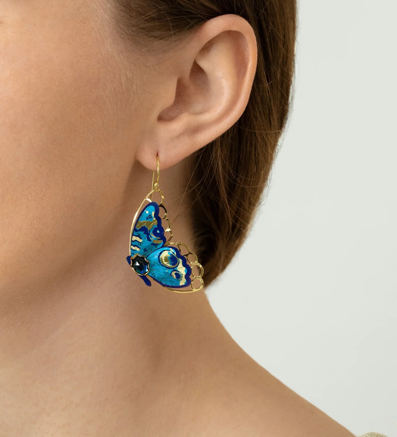 A close-up of a person wearing Holly Yashi’s Mariposa Butterfly Wing Blue Radiance Earrings, shaped like blue butterflies with gold accents. Their hair is tucked behind their ear, and they are dressed in a light knit top.