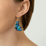A close-up of a person wearing Holly Yashi’s Mariposa Butterfly Wing Blue Radiance Earrings, shaped like blue butterflies with gold accents. Their hair is tucked behind their ear, and they are dressed in a light knit top.