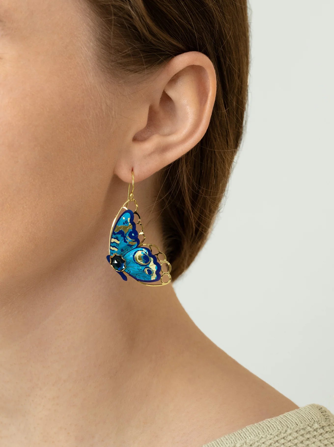 A close-up of a person wearing Holly Yashi’s Mariposa Butterfly Wing Blue Radiance Earrings, shaped like blue butterflies with gold accents. Their hair is tucked behind their ear, and they are dressed in a light knit top.