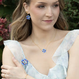 A woman in a light, ruffled dress poses outdoors, accessorized with Holly Yashi’s Blissful Bloom Double Strand Bonnie Blue Bracelet and blue flower jewelry. Pink flowers and green foliage complete the delicate scene.