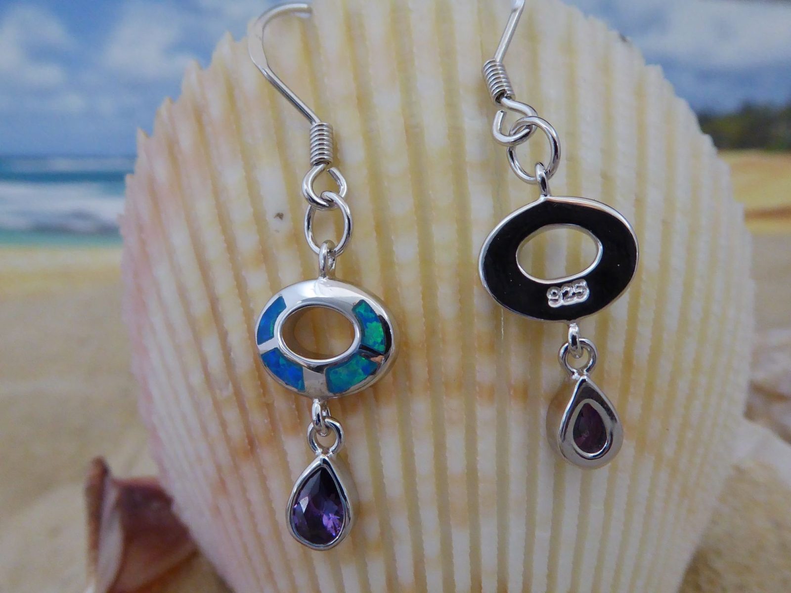 Jewelry Network’s Caribbean Opal Oval w/Amethyst Teardrop Dangle Earrings are shown before a seashell, with the beach and ocean softly blurred in the background.