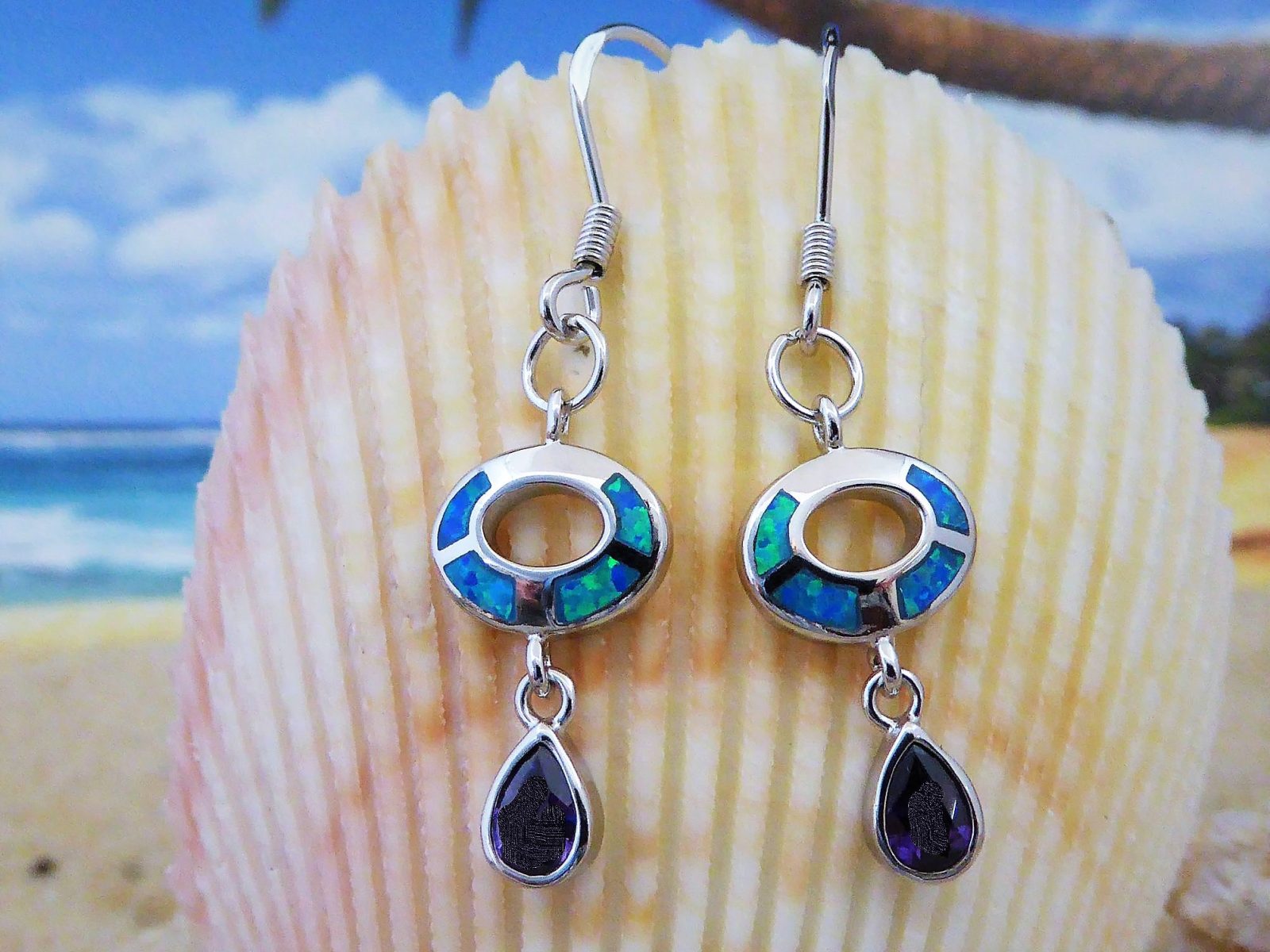 Jewelry Network's Caribbean Opal Oval w/Amethyst Teardrop Dangle Earrings are shown before a large seashell, with a beach scene in the background.