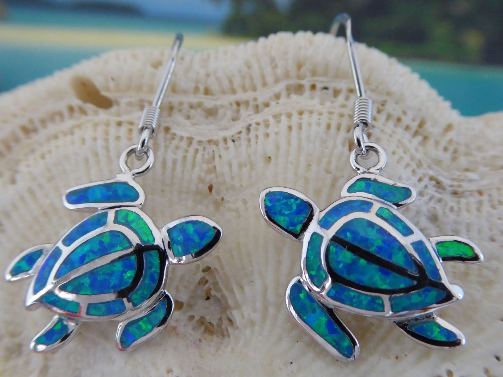 Jewelry Network’s Caribbean Opal Turtle/Full Shell Dangle Earrings feature silver sea turtles with blue and green synthetic opal inlay, hanging from hooks and shown against a textured seashell background.