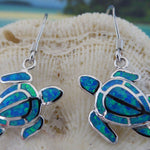 Jewelry Network’s Caribbean Opal Turtle/Full Shell Dangle Earrings feature silver sea turtles with blue and green synthetic opal inlay, hanging from hooks and shown against a textured seashell background.
