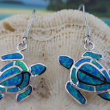 Jewelry Network’s Caribbean Opal Turtle/Full Shell Dangle Earrings feature silver sea turtles with blue and green synthetic opal inlay, hanging from hooks and shown against a textured seashell background.