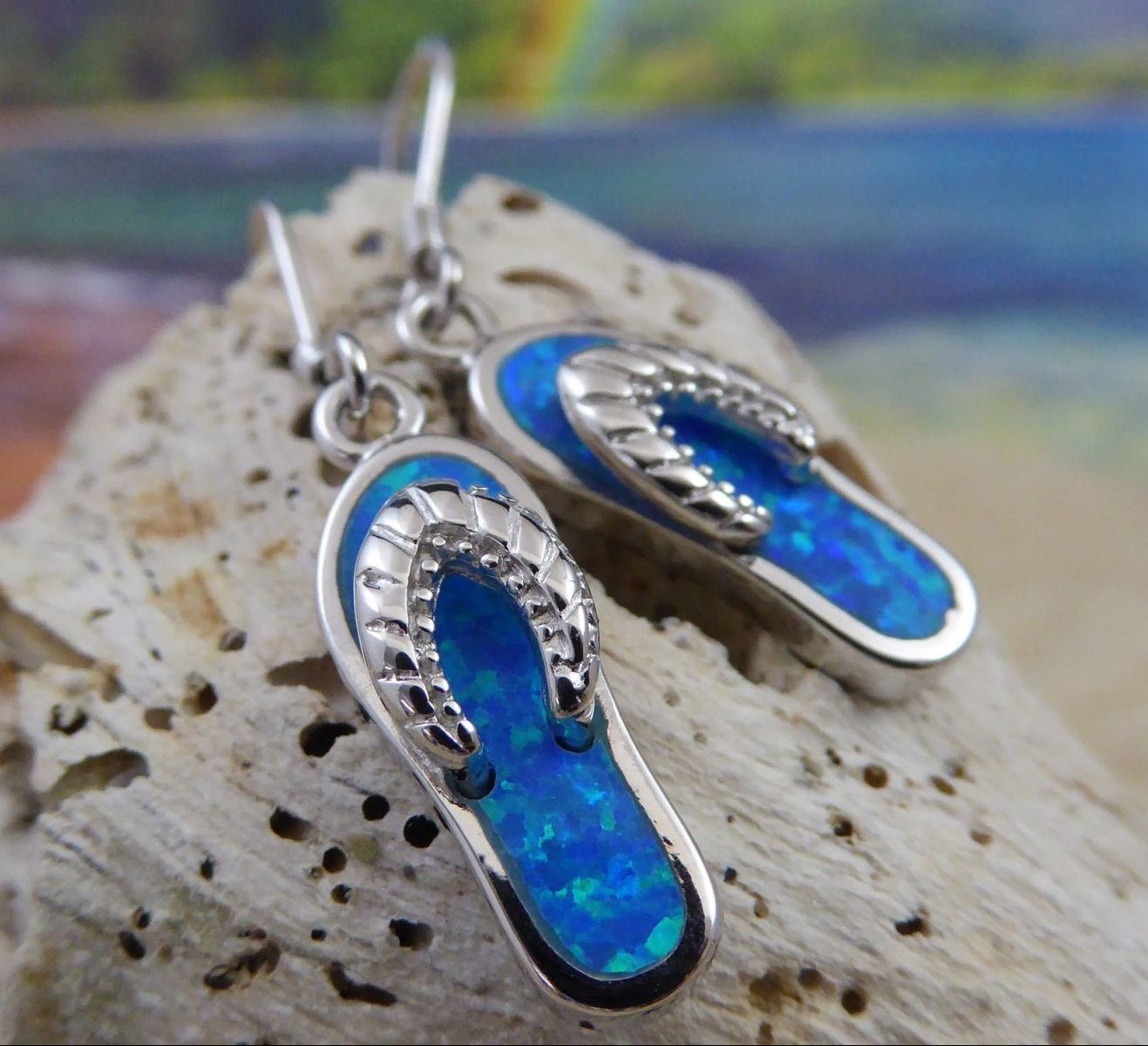 The Caribbean Opal Flip Flop Dangle Earrings by Jewelry Network feature blue opal inlays in 925 sterling silver, displayed on a textured rock with a vibrant, beach-inspired background—ideal for seashell jewelry lovers.