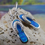 The Caribbean Opal Flip Flop Dangle Earrings by Jewelry Network feature blue opal inlays in 925 sterling silver, displayed on a textured rock with a vibrant, beach-inspired background—ideal for seashell jewelry lovers.