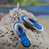 The Caribbean Opal Flip Flop Dangle Earrings by Jewelry Network feature blue opal inlays in 925 sterling silver, displayed on a textured rock with a vibrant, beach-inspired background—ideal for seashell jewelry lovers.
