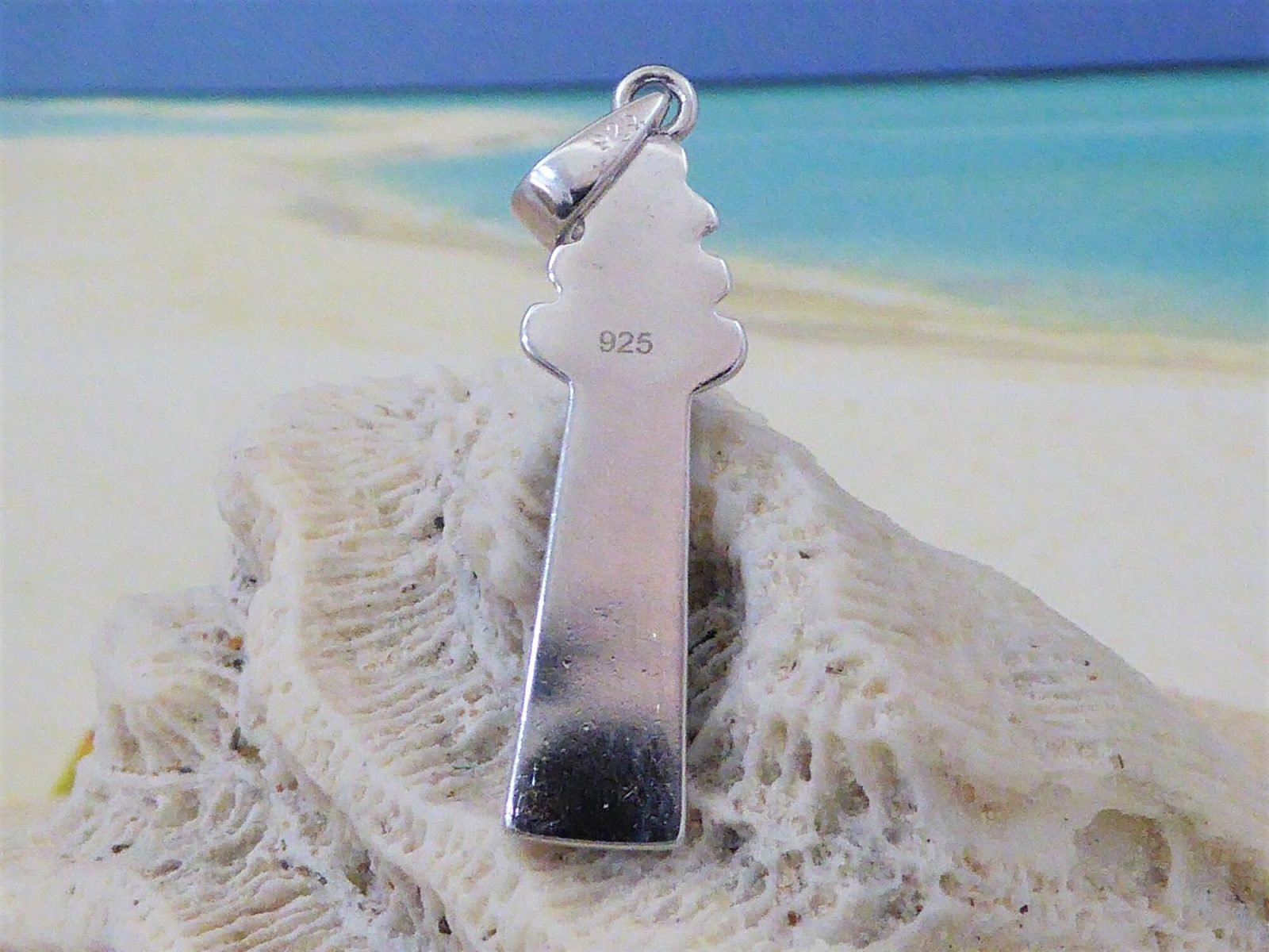 The Caribbean Opal Lighthouse-Pendant ONLY by Jewelry Network, crafted in sterling silver and marked 925, rests on textured white coral with a sandy beach and blue ocean blurred in the background—a striking nautical accessory.