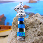 The Jewelry Network Caribbean Opal Lighthouse-Pendant ONLY features alternating blue and black striped inlays in sterling silver, displayed on a textured rock with a blurred seascape and rocks in the background—a stunning nautical piece.