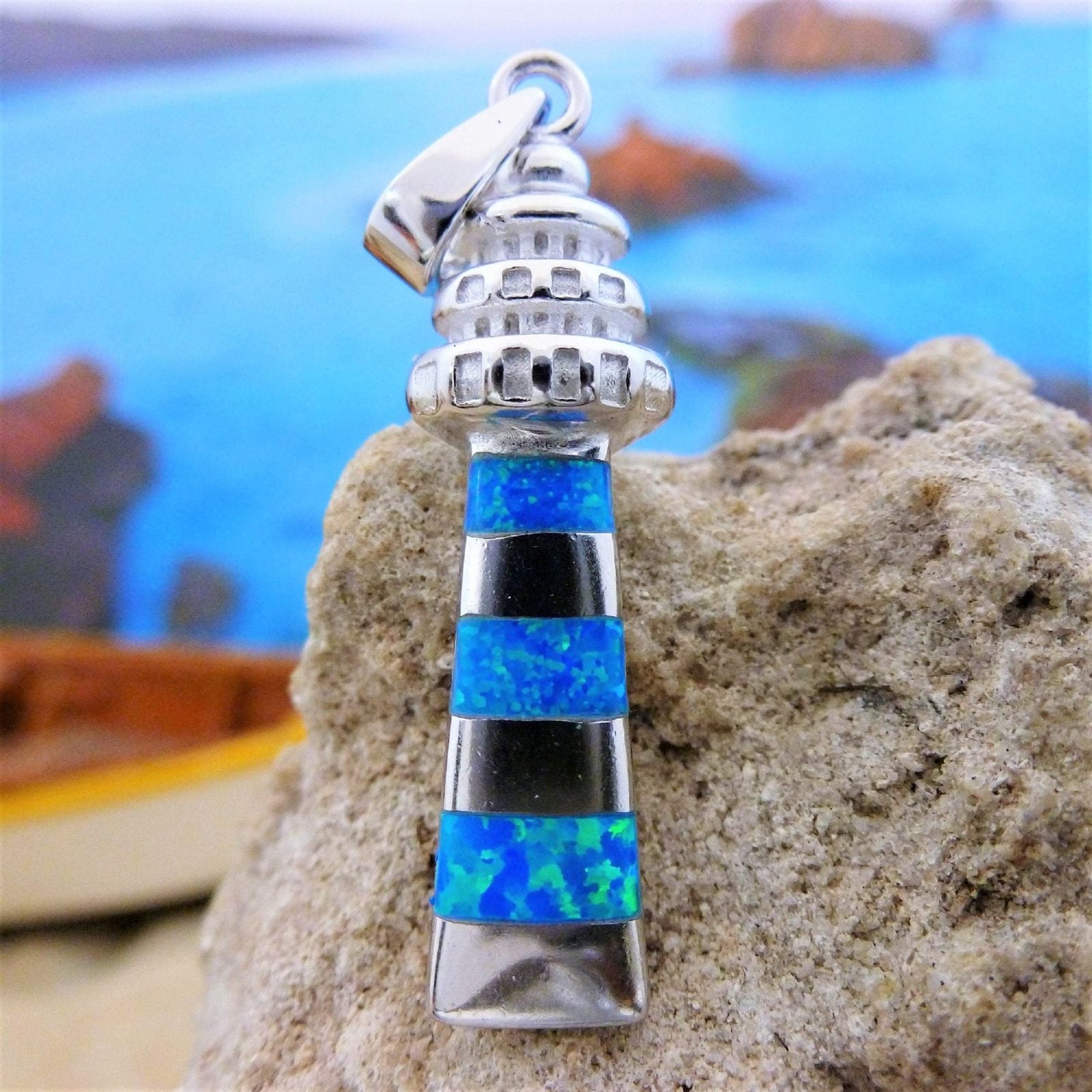 The Jewelry Network Caribbean Opal Lighthouse-Pendant ONLY features alternating blue and black striped inlays in sterling silver, displayed on a textured rock with a blurred seascape and rocks in the background—a stunning nautical piece.
