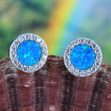 Jewelry Network's Caribbean Opal Round w/CZ Surround Post Earrings feature vibrant blue synthetic opal centers encircled by sparkling cubic zirconia, elegantly showcased on a textured brown seashell with a colorful blurred background.