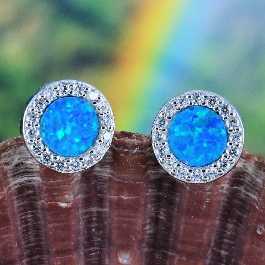 Jewelry Network's Caribbean Opal Round w/CZ Surround Post Earrings feature vibrant blue synthetic opal centers encircled by sparkling cubic zirconia, elegantly showcased on a textured brown seashell with a colorful blurred background.