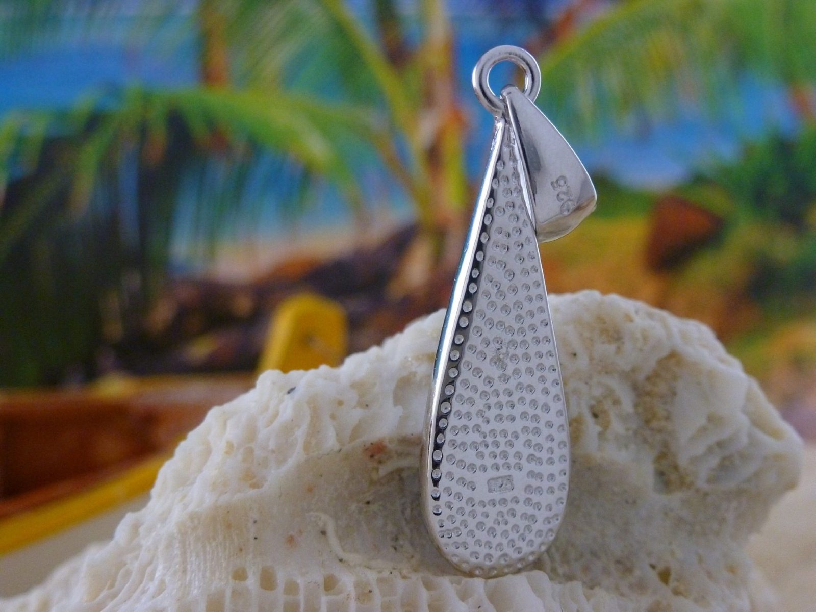 The Jewelry Network Caribbean Opal Teardrop 4 Segments pendant rests on coral, with a blurred tropical background of palm trees and blue sky.