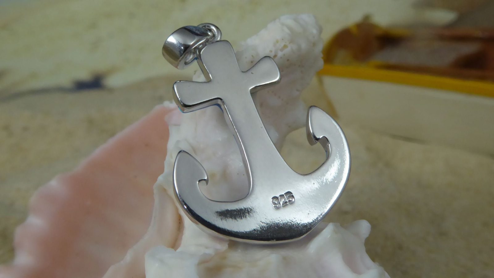 The Caribbean Opal Anchor w/ Segments-Pendant ONLY by Jewelry Network, a shiny silver 925 Sterling Silver anchor engraved with "925," sits on a white seashell with sand and a blurred yellow object in the background.