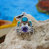 The Jewelry Network Caribbean Opal Crab w/Round Amethyst CZ-Pendant rests on a rough beige rock, with its silver finish and blue-green synthetic opal accents shining against a blurred blue and brown background.
