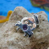 A Caribbean Opal Crab pendant with a round Amethyst CZ by Jewelry Network, stamped 925, rests on a textured tan rock. The background is blurred with blue and orange hues. Pendant only; chain not included.