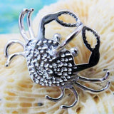 A close-up of the Jewelry Network Caribbean Opal "Heart" Shaped Crab-Pendant ONLY, crafted in shiny 925 Sterling Silver with detailed legs and pincers, displayed on a textured beige surface against a blurred blue-green background.
