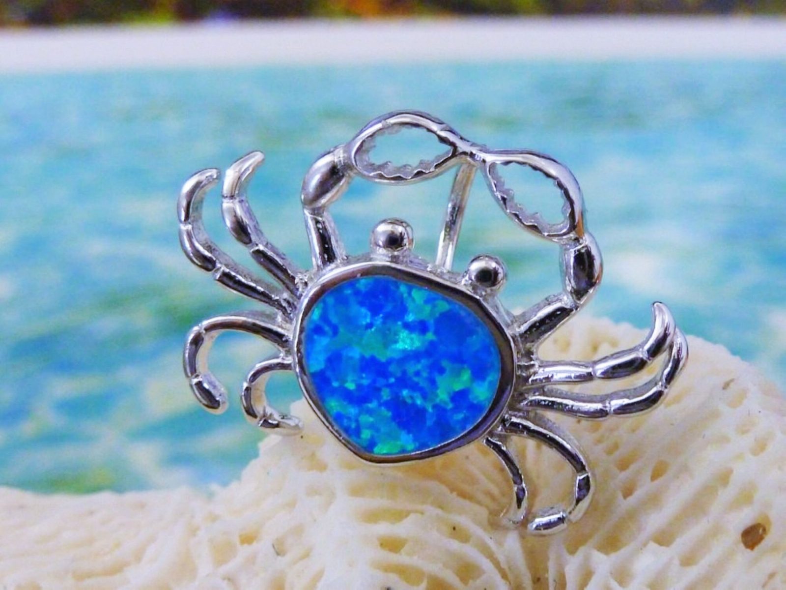 The Jewelry Network Caribbean Opal "Heart" Shaped Crab-Pendant features a silver crab design with a vivid synthetic opal center, displayed on white coral against a backdrop of turquoise ocean and sandy beach.