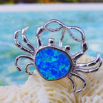 The Jewelry Network Caribbean Opal "Heart" Shaped Crab-Pendant features a silver crab design with a vivid synthetic opal center, displayed on white coral against a backdrop of turquoise ocean and sandy beach.