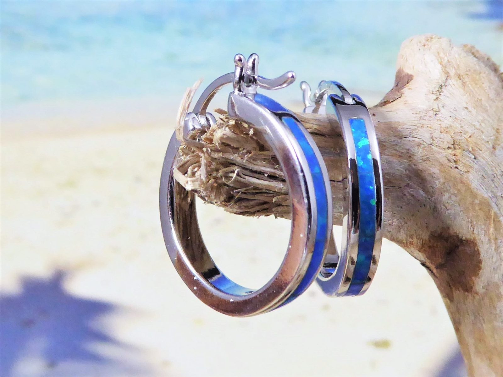 The Jewelry Network Caribbean Opal Solid Small Hoop 20MM earring, featuring a blue opal inlay, hangs on driftwood with a blurred sandy beach and ocean in the background.