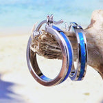 The Jewelry Network Caribbean Opal Solid Small Hoop 20MM earring, featuring a blue opal inlay, hangs on driftwood with a blurred sandy beach and ocean in the background.