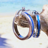 The Jewelry Network Caribbean Opal Solid Small Hoop 20MM earring, featuring a blue opal inlay, hangs on driftwood with a blurred sandy beach and ocean in the background.