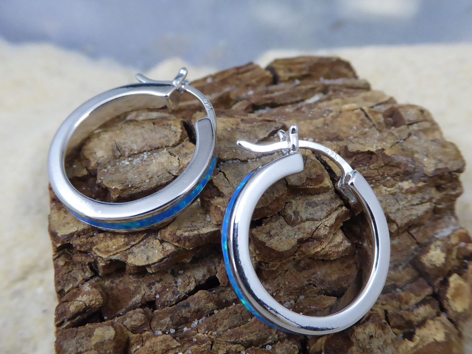 The Caribbean Opal Solid Small Hoop 20MM from Jewelry Network, crafted in sterling silver with a blue accent, is displayed on textured brown wood against a blurred backdrop.