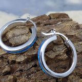 The Caribbean Opal Solid Small Hoop 20MM from Jewelry Network, crafted in sterling silver with a blue accent, is displayed on textured brown wood against a blurred backdrop.