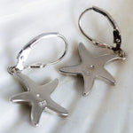 Jewelry Network's Caribbean Opal Starfish Dangle Earrings are crafted from 925 Sterling Silver with rhodium plating and leverback hooks, featuring starfish shapes and engraved markings on the backs, displayed on a white surface.