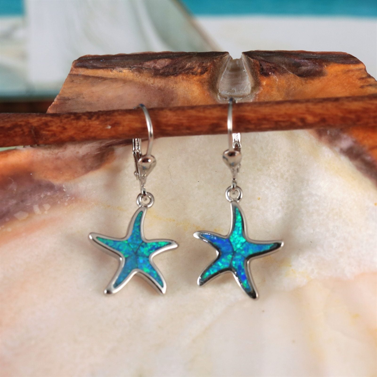 Jewelry Network's Caribbean Opal Starfish Dangle Earrings feature 925 Sterling Silver with rhodium plating, blue and green synthetic opal centers, and hook backs, displayed against a large seashell.