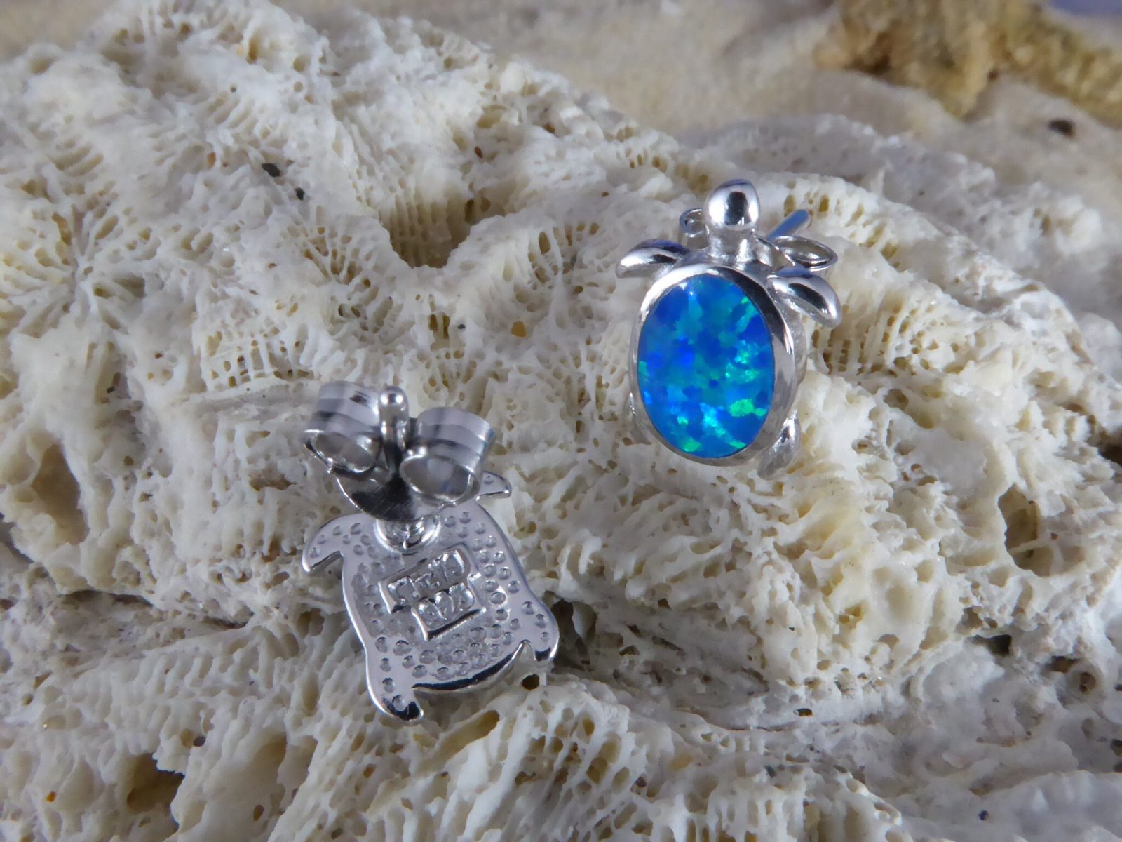 Two Caribbean Opal Turtle Stud Earrings by Jewelry Network, each rhodium plated with a 925 hallmark, rest on textured coral—one displays a bright blue-green opal shell, the other shows its finely engraved back.