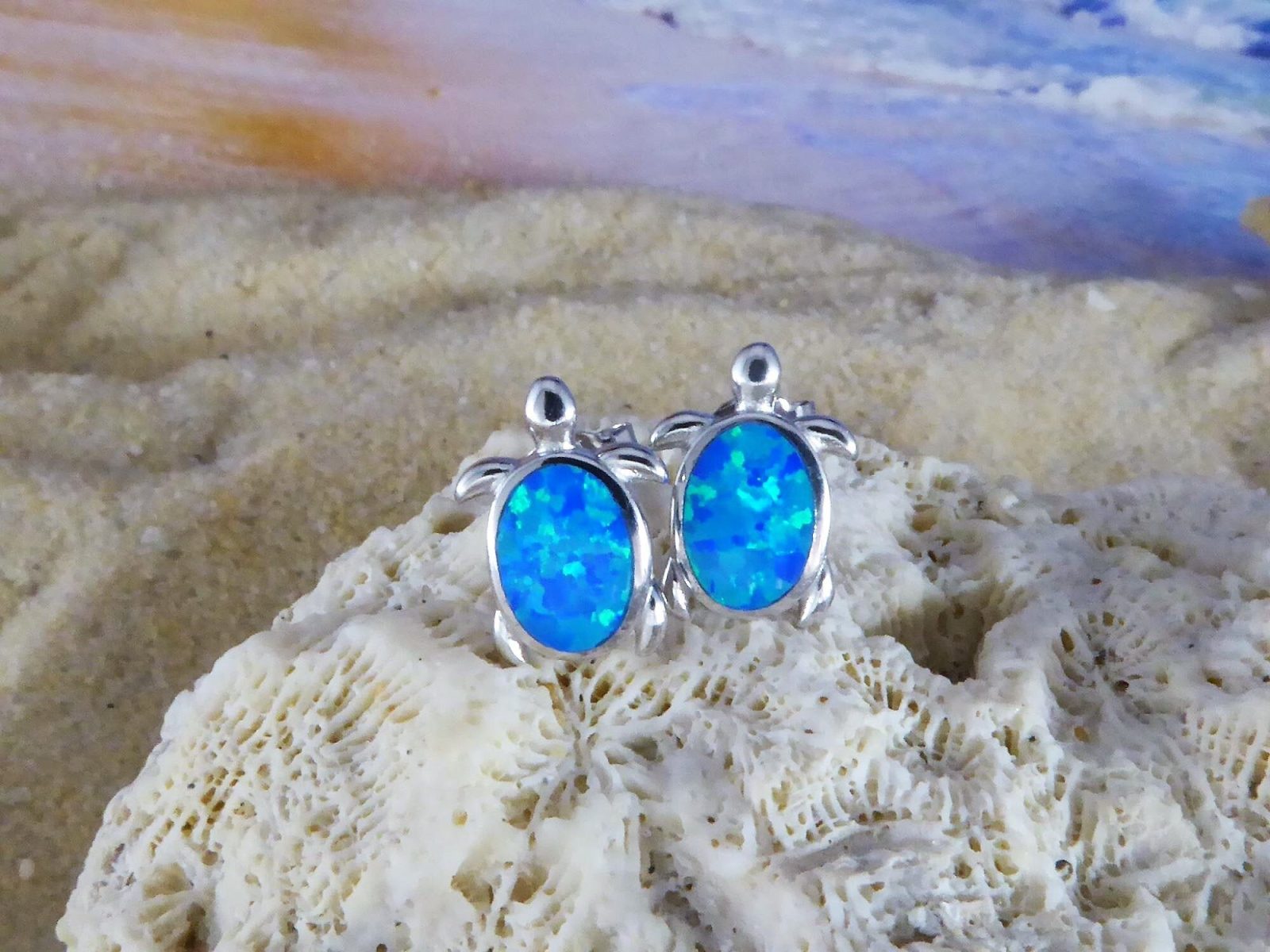 The Jewelry Network Caribbean Opal Turtle Stud Earrings feature silver turtle shapes with bright blue opal-like stones and a rhodium plated finish, shown on coral against a blurred sandy beach and ocean backdrop.