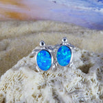 The Jewelry Network Caribbean Opal Turtle Stud Earrings feature silver turtle shapes with bright blue opal-like stones and a rhodium plated finish, shown on coral against a blurred sandy beach and ocean backdrop.